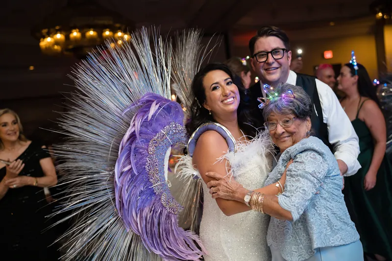 Lehna in a white dress stands with a man in glasses and an older woman holding a large purple feathered fan prop.