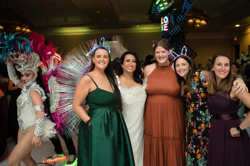 Five women in various colored dresses stand together smiling, with elaborately costumed performers visible to their left