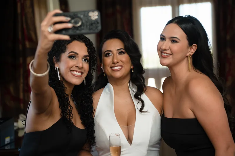 Three women take a selfie together indoors, with the center figure wearing Lehna's white dress.