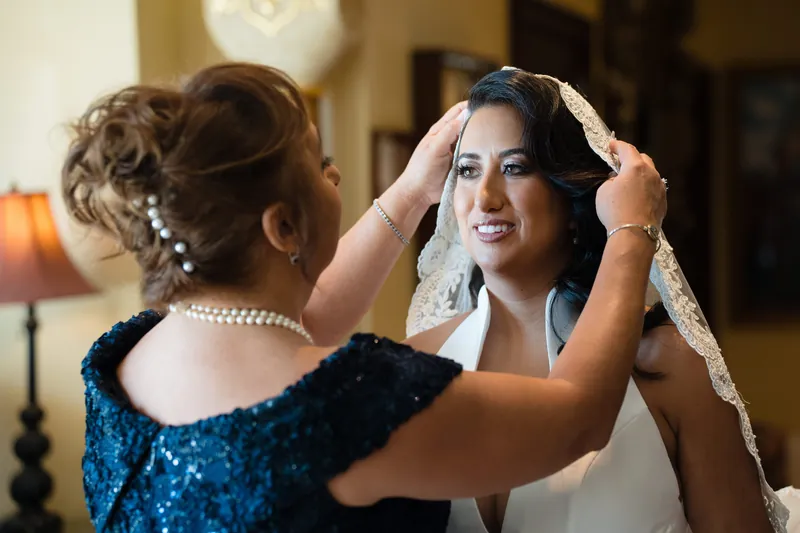 A woman adjusts Lehna's veil and headpiece indoors as Lehna smiles, both wearing formal attire.