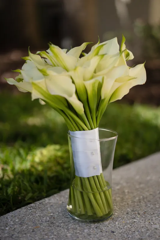 Yellow and white calla lilies bundled with greenery sit in a clear glass vase on stone ground.