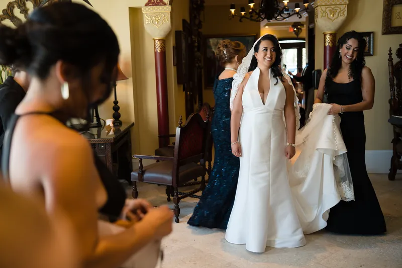 Lehna walks through an ornate interior hallway in a white wedding dress, surrounded by guests in formal wear.
