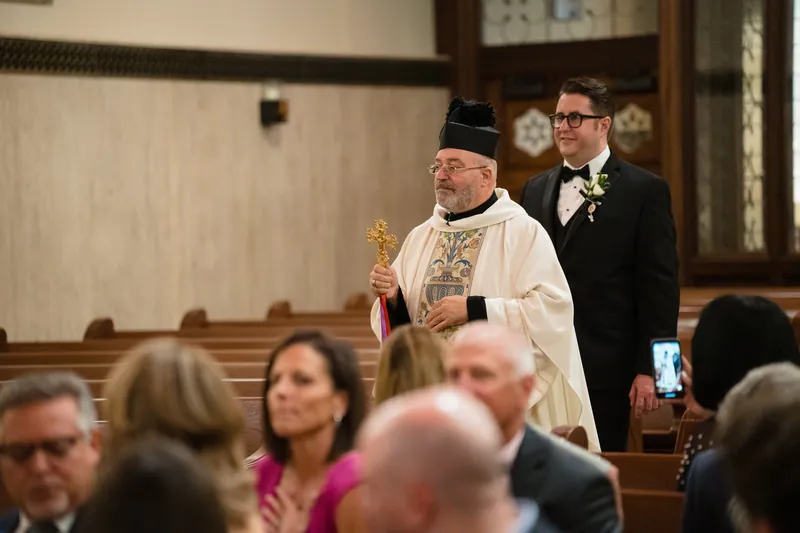 A clergy member in white vestments stands at the altar during the ceremony as Cole watches from nearby.