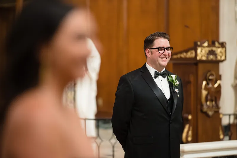 Cole waits at the altar in a black tuxedo and glasses, watching as Lehna approaches down the aisle.