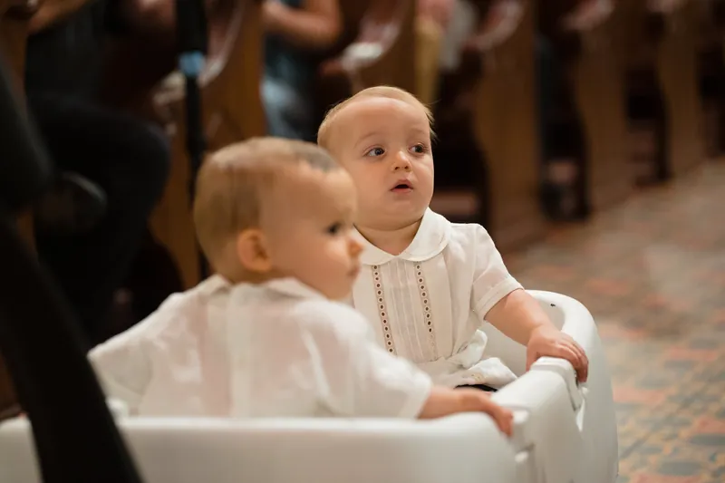 Two young children in white christening gowns sit together in the pews during the ceremony at the basilica.