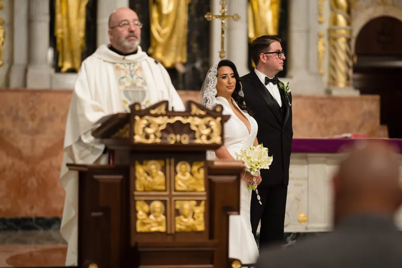 A priest in white vestments stands at the altar beside Cole and Lehna during their ceremony at the Cathedral Basilica of St.