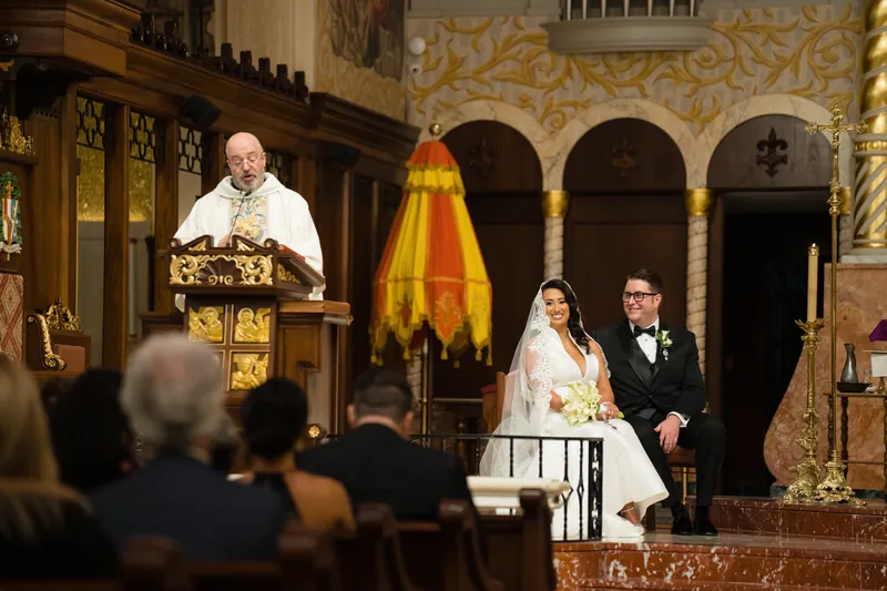 Priest at altar in golden vestments addresses Lehna and Cole during their ceremony at the Cathedral Basilica of St.