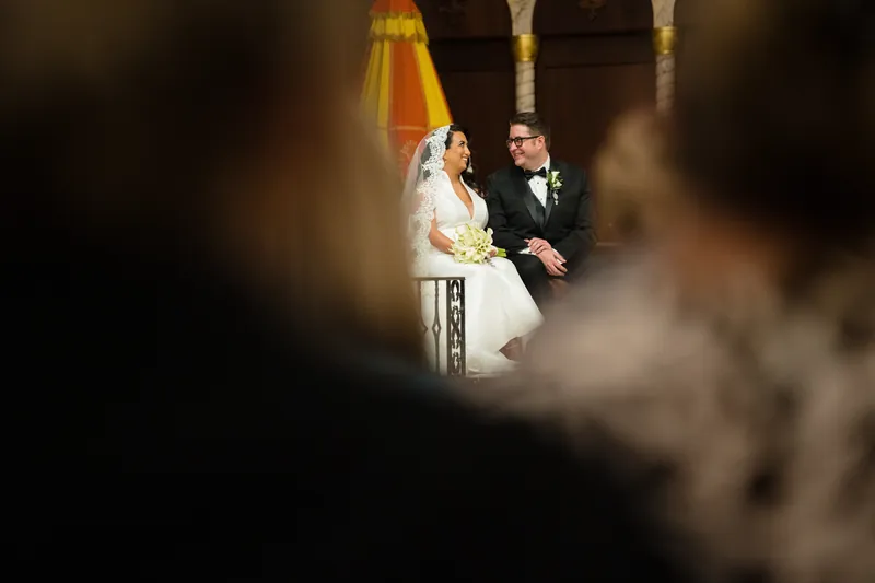 Lehna and Cole stand together at the altar, framed by guests' shoulders in foreground and ornate architecture behind.