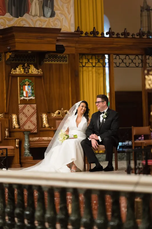 Lehna in white wedding dress and Cole in black suit sit together in wooden pews during their ceremony.