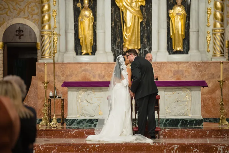 Cole kisses Lehna at the altar, her white gown and veil contrasting against gilded saint statues and marble details behind