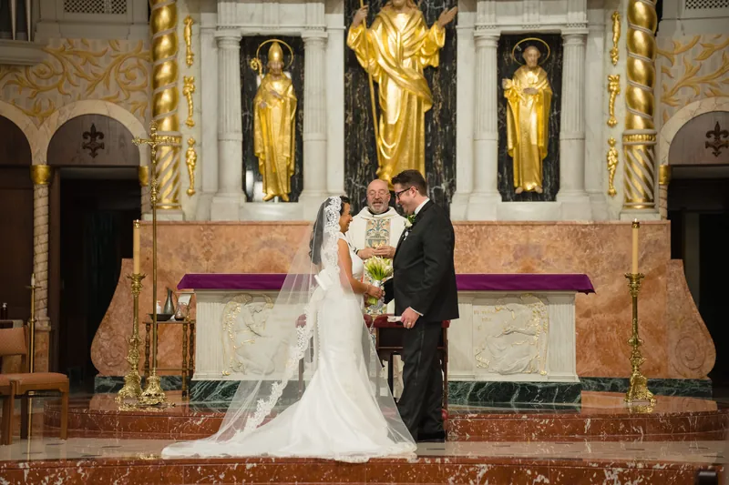 Lehna and Cole hold hands and smile while standing before the altar, surrounded by candlelight and golden religious