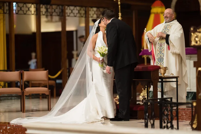 A clergy member in white vestments presides as Lehna and Cole share a moment at the altar, her flowing veil trailing across