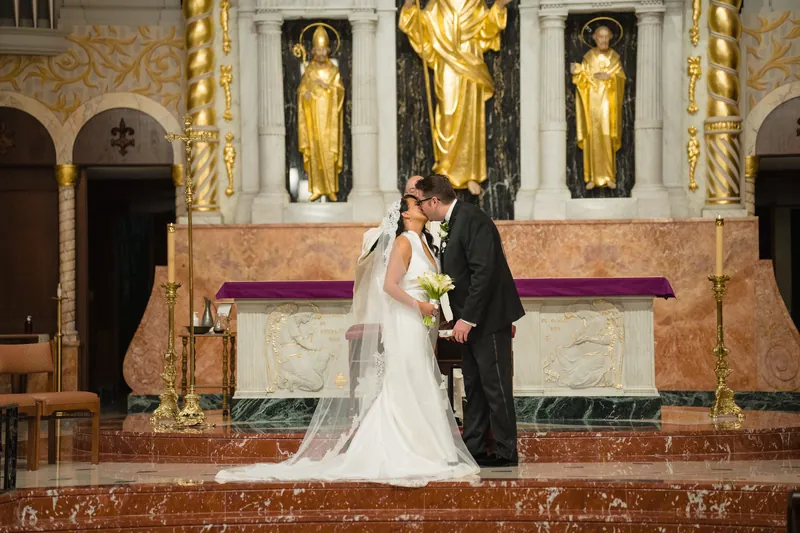 Cole dips Lehna in a kiss at the altar, her white dress and veil creating movement against the ornate architectural backdrop.