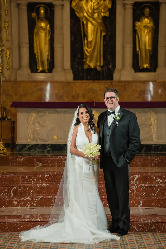 Lehna in a white wedding gown and Cole in a dark suit stand together at the Cathedral Basilica's altar, golden saint statues