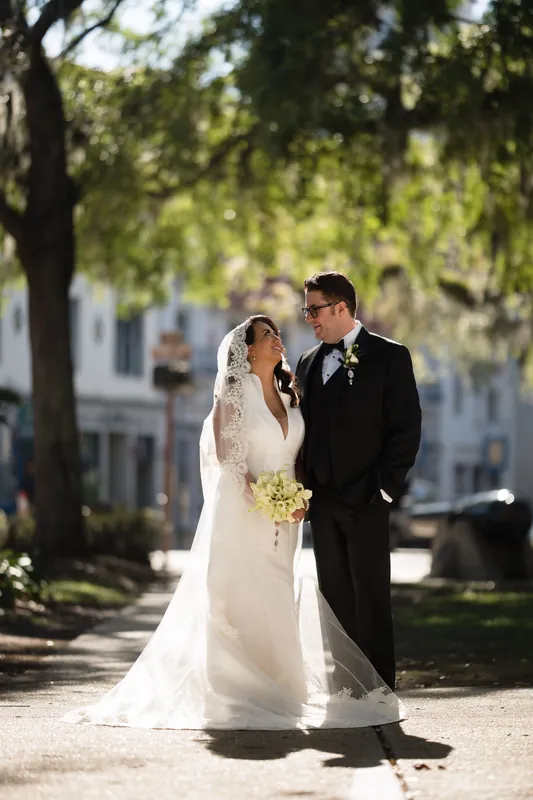 Lehna and Cole walk together through a tree-lined courtyard at Casa Monica, she in her white lace gown and veil, he