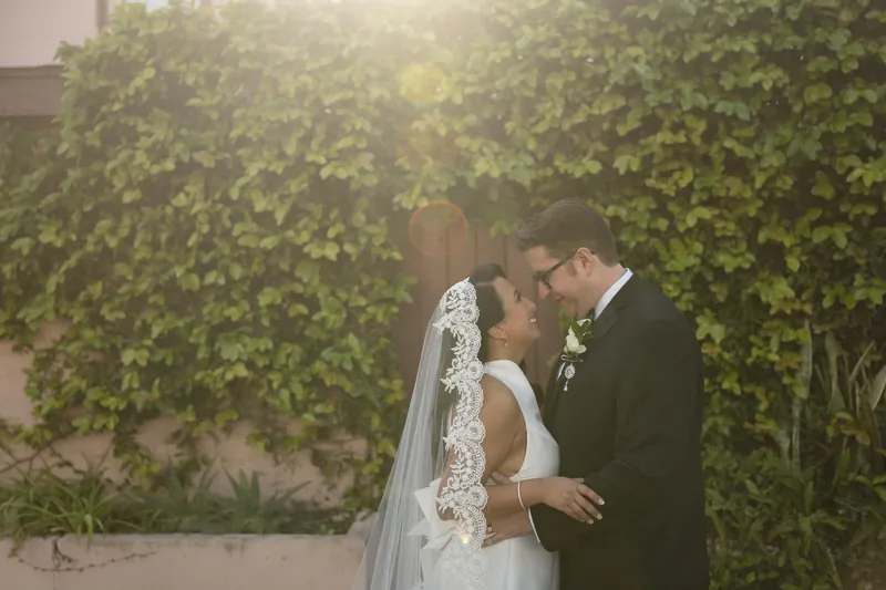 Cole leans in to kiss Lehna against the lush ivy wall, her lace veil visible as they embrace.
