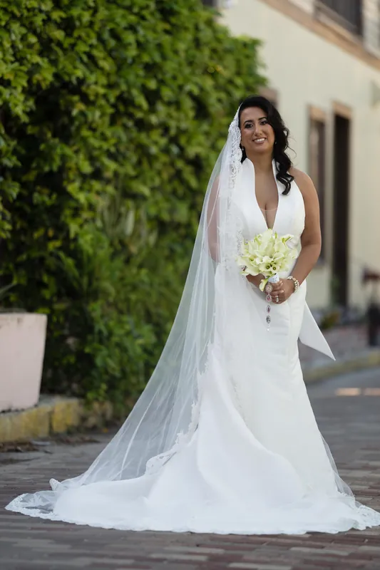 Lehna stands alone on the resort's patio in her sleeveless white gown, holding a white bouquet with her veil trailing behind.