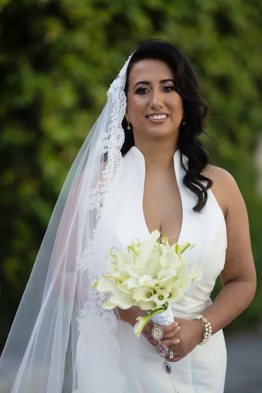 Lehna smiles at the camera holding her white floral bouquet, her long veil draped over one shoulder against green foliage.