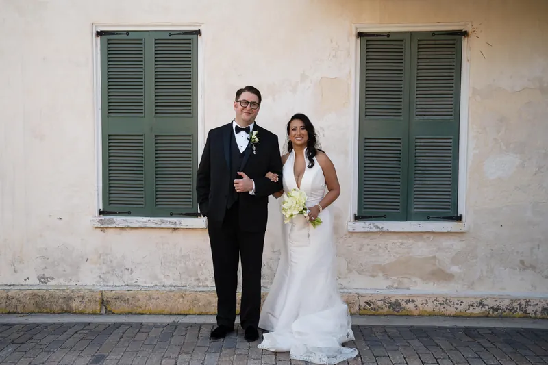 Cole and Lehna stand arm-in-arm on a brick pathway between whitewashed walls with green shutters.