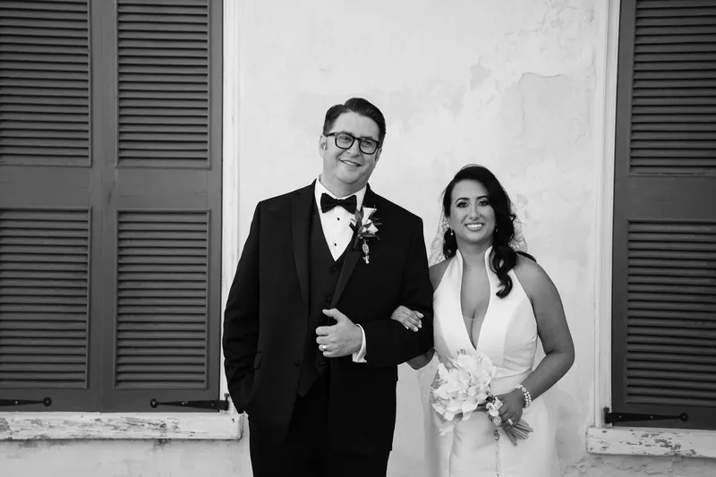 Black and white shot: Cole and Lehna walk together in formal wedding attire past the resort's colonial-style shuttered doors.