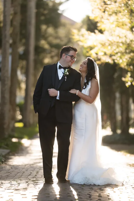 Cole and Lehna walk down a tree-lined brick path, sunlight filtering through the leaves as they stroll together.