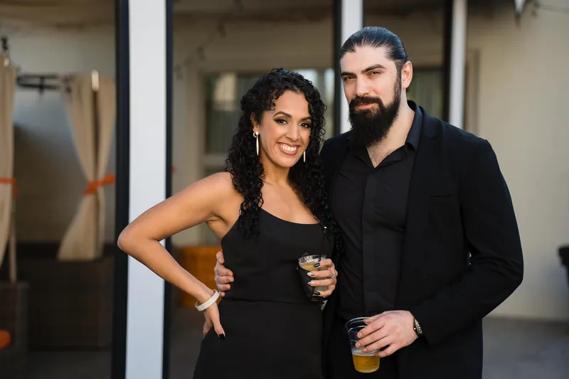 Two guests in black formal wear stand together holding drinks indoors.
