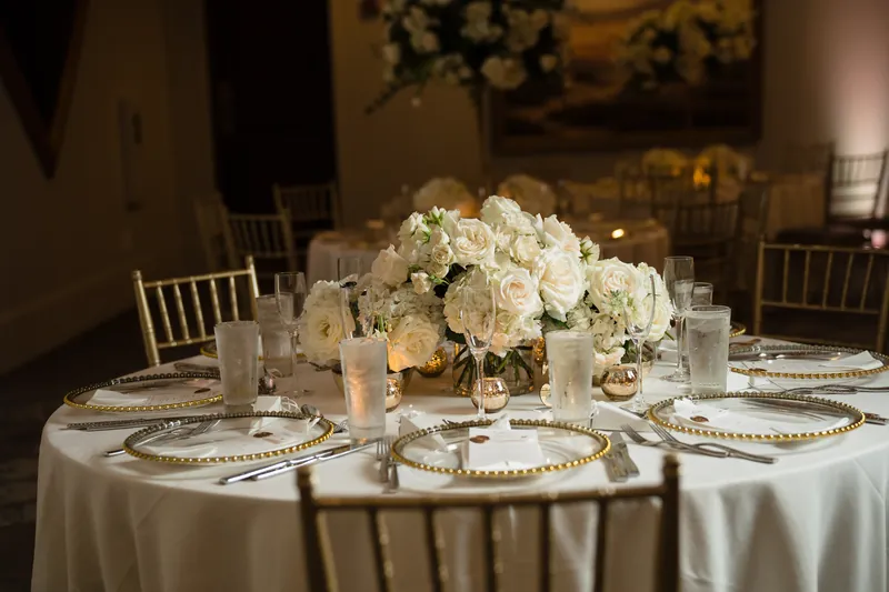 White roses and hydrangeas fill a tall centerpiece above a round table set with gold-rimmed plates and napkins.