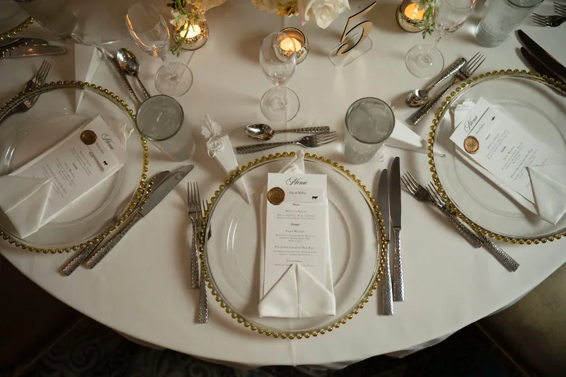 Three place settings feature white plates on gold charger rings with rope detailing and menu cards at a round table.