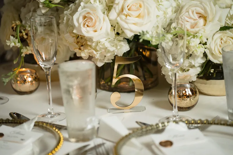 Cream and white roses with baby's breath fill a low arrangement next to champagne flutes and gold accents on the table.