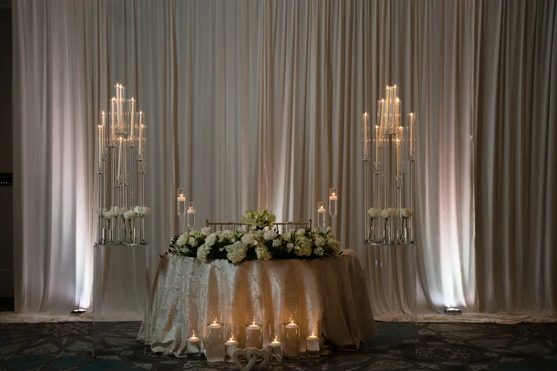 Two tall brass candelabras flank a white floral arrangement on a draped table with gray curtains and uplighting behind it.