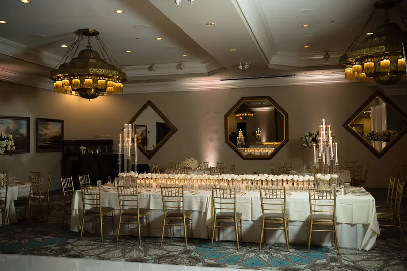 Four long banquet tables with white linens and gold chiavari chairs fill the elegant ballroom beneath ornate chandeliers