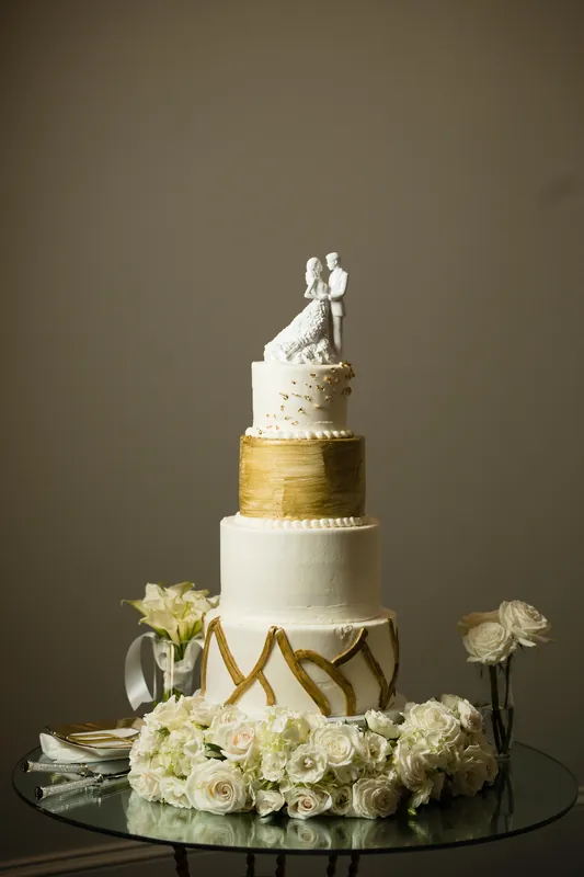 Multi-tiered wedding cake with white fondant, gold geometric details, and a couple topper sits surrounded by cream-colored