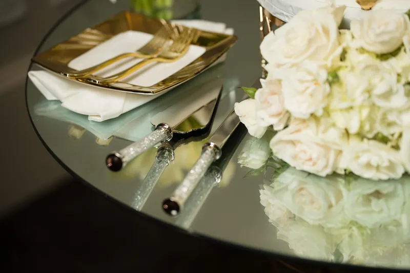 Decorative cake knife and server with jeweled handles rest on a glass table next to white floral arrangements