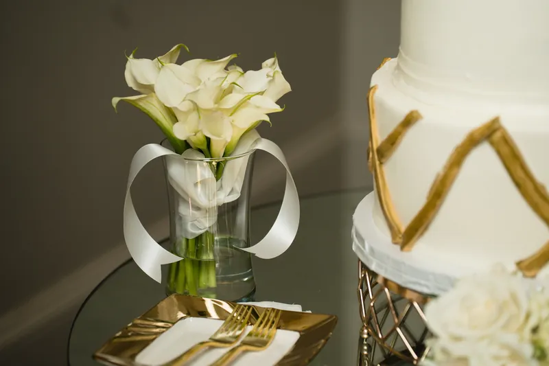 White calla lilies in a clear vase stand beside tiered cake layers featuring gold trim and brushstroke details.