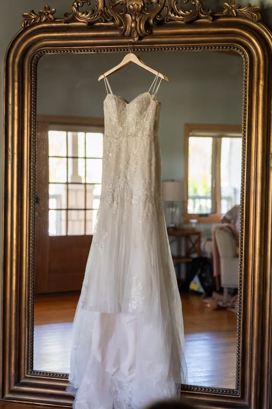Lena's lace wedding gown hangs from a gold-framed mirror, its train pooling on the wooden floor.