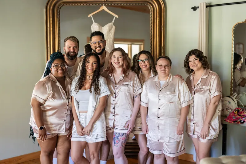 Lena stands with her bridal party in matching satin robes, gathered in front of the gold-framed mirror.