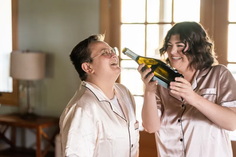 Two bridesmaids playfully share a champagne bottle while getting ready, both in matching robes.