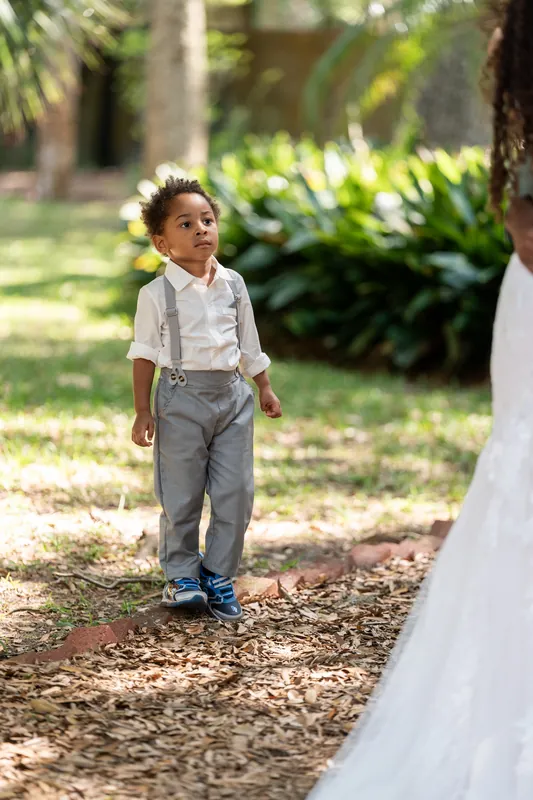 A small boy in suspenders and white shirt watches Lena's white gown trail past him on the woodland grounds.