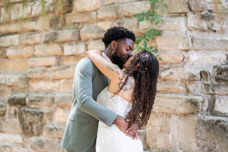 Blake and Lena embrace against a weathered stone wall, she in her white lace gown and he in a gray suit with gold vest.