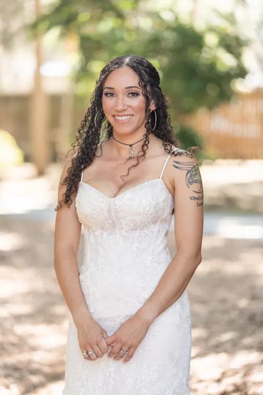 Lena smiles at the camera in her beaded white wedding dress, standing alone on a tree-lined path at Fountain of Youth