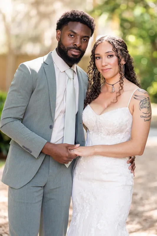 Blake and Lena stand together on a shaded pathway, holding hands and gazing toward the camera in their wedding attire.