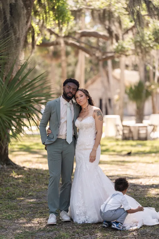 Blake and Lena stand together under moss-draped trees at Fountain of Youth Archaeological Park, with a young child sitting