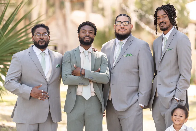Blake stands with three groomsmen in matching gray suits among the historic grounds, all posed confidently with a young child