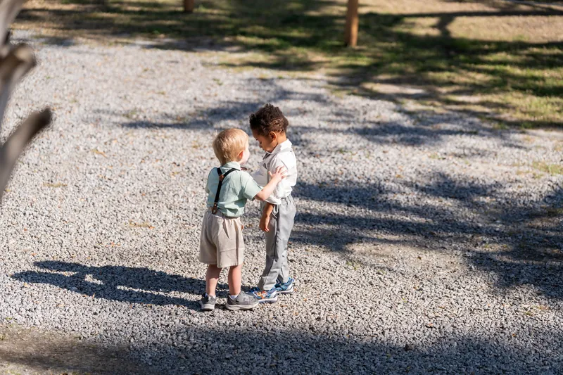 Two small children hold hands and walk together across the sandy grounds scattered with tree shadows at Fountain of Youth