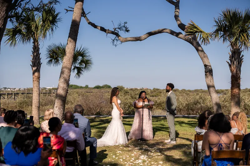 Guests seated on blue chairs witness Lena and Blake's wedding ceremony framed by a dramatic natural arch of intertwined