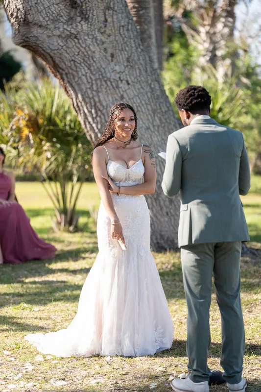 Lena stands in her flowing white gown beside Blake beneath a gnarled oak tree, surrounded by guests in the grassy venue.
