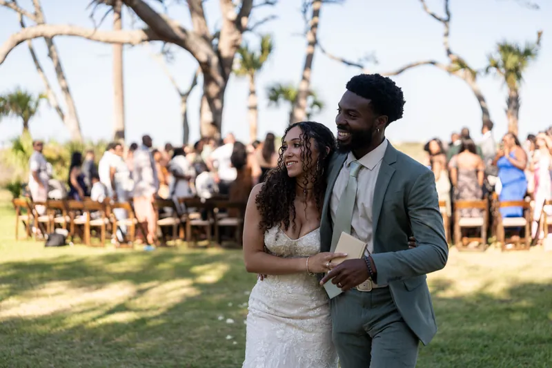 Blake stands beside Lena under oak trees at Fountain of Youth Archaeological Park, both smiling as wedding guests gather