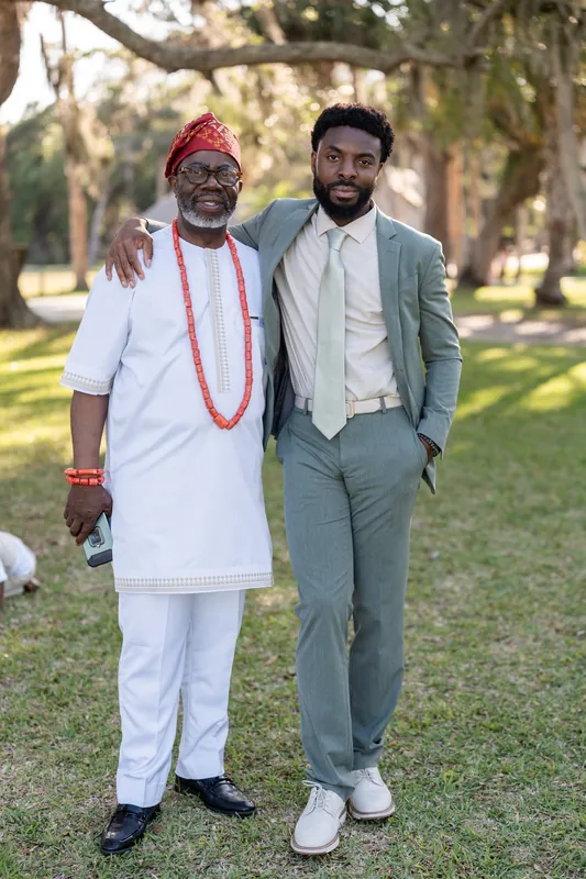 Blake's father in white traditional attire and red beads stands arm-in-arm with Blake in his gray suit on the park's lawn.