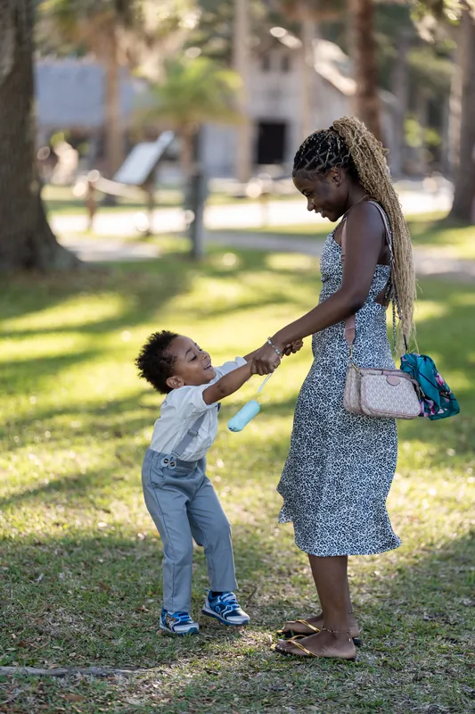 A young child reaches up to hold hands with a woman in a blue dress on the grass beneath ancient oak trees.