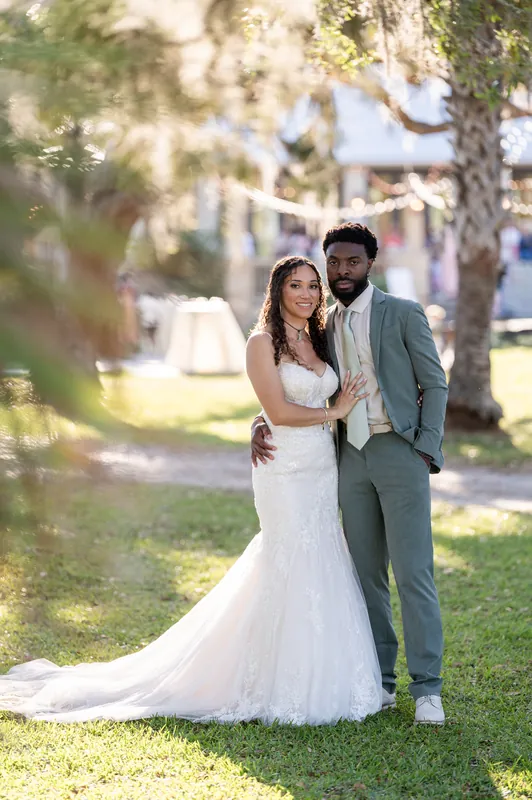 Lena wears a flowing white mermaid gown while Blake stands beside her in a sage green suit under oak trees at Fountain
